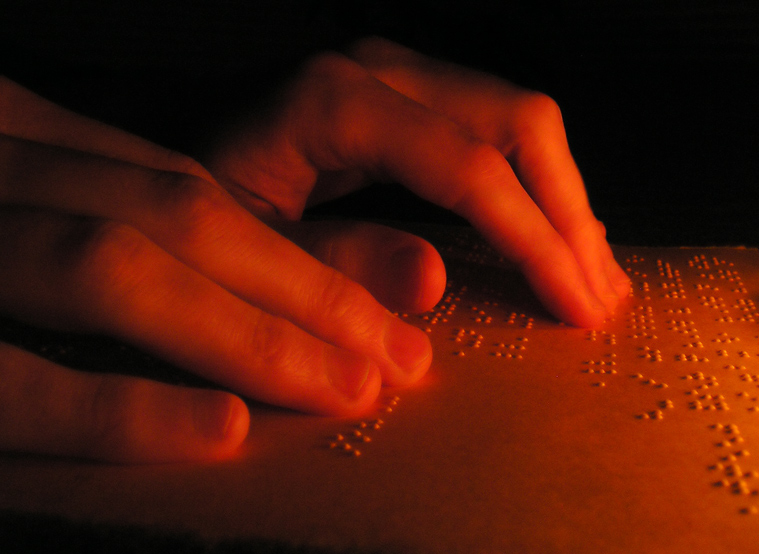 Image of 2 hands reading braille, hands are seen from the side. The whole image in low light with black and various tones of orange and fiery red Braille dots cast small gentle shadows.  THe written Braille is a partial grocery list and quote form the book Lord of the rings.
